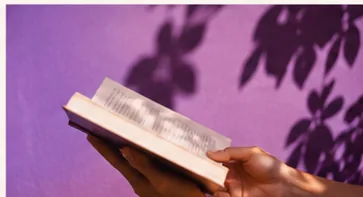 Livro aberto em mãos diante de fundo lilás